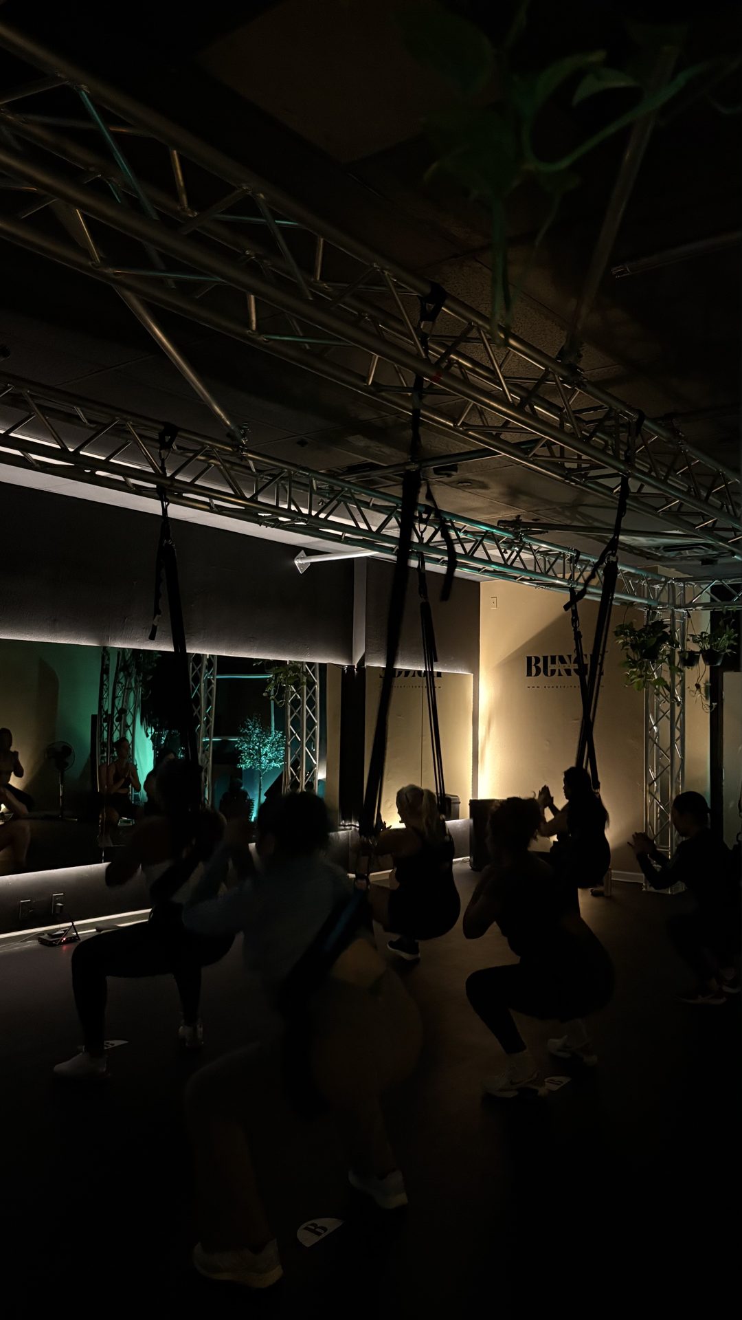 A group squat sequence under teal lighting during a bungee fitness class at BUNGE Fitness